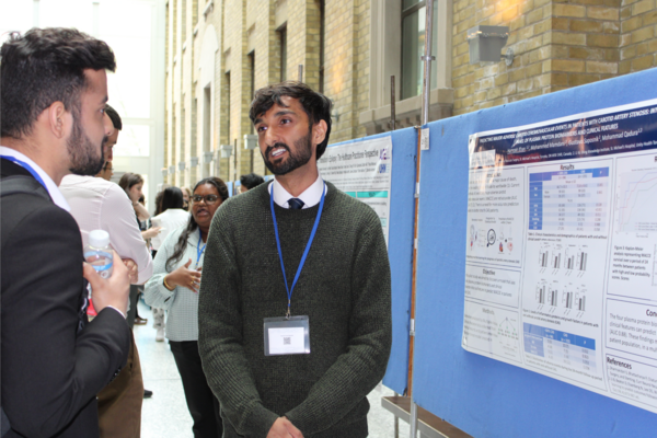 Hamza Khan stands in front of his research poster, speaking with another person. 